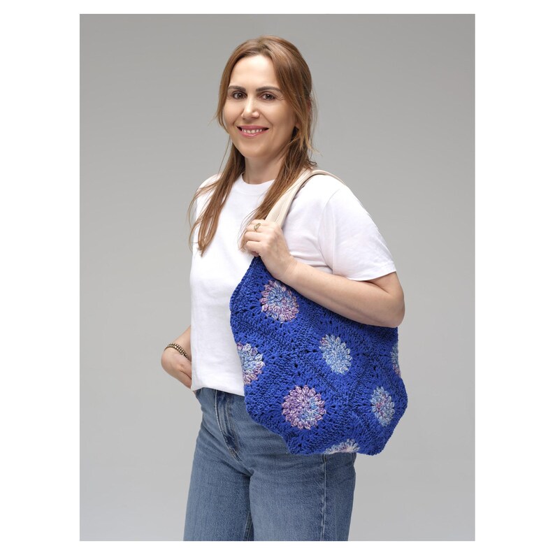 May include: A blue crochet tote bag with a floral pattern. The bag features a series of crocheted flower motifs in shades of blue, purple, and pink. The bag has a cream-colored strap and is held by a person wearing a white t-shirt and blue jeans.