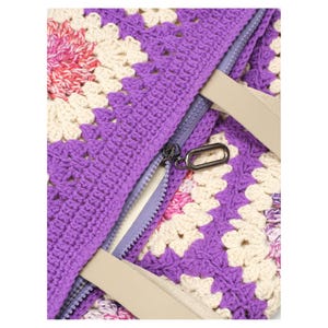 May include: Close-up of a crocheted tote bag with a vibrant floral pattern in purple, cream, pink, and white. The bag features a zipper closure and beige straps. The crochet stitches are clearly visible, showcasing the handmade quality.