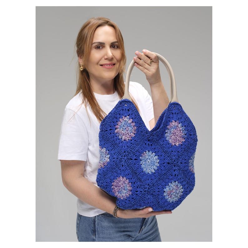 May include: A blue crocheted tote bag with a floral pattern in shades of blue, purple, and pink. The bag has cream-colored handles and is held up by a person. The bag is a stylish accessory.