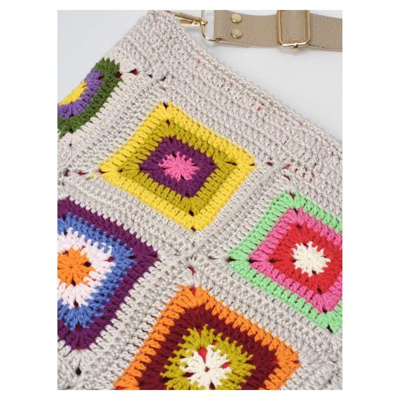 May include: Close-up of a crocheted bag with a neutral grey base. The bag features colourful, square granny squares in shades of yellow, purple, green, orange, and blue. A beige strap with gold-coloured hardware is attached to the bag.