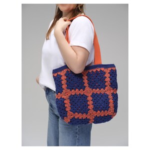 May include: A blue and orange crochet tote bag with a geometric pattern. The bag has a solid orange strap and is carried over the shoulder. The bag is made of yarn and has a handmade appearance.