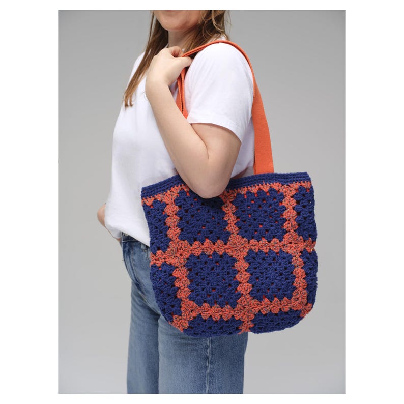 May include: A blue and orange crochet tote bag with a geometric pattern. The bag has a solid orange strap and is carried over the shoulder. The bag is made of yarn and has a handmade appearance.