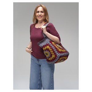 May include: A crochet tote bag featuring a granny square pattern in shades of burgundy, mustard yellow, and grey. The bag has a wide grey shoulder strap and a structured, boxy shape, constructed from yarn with a visible textured stitch pattern.