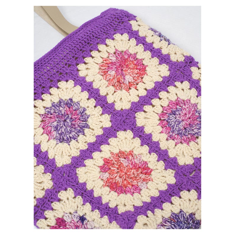 May include: A handmade crochet tote bag featuring a vibrant granny square pattern. The bag showcases a purple border and handle, with cream, pink, and purple floral-like squares. The bag is a stylish and functional accessory.