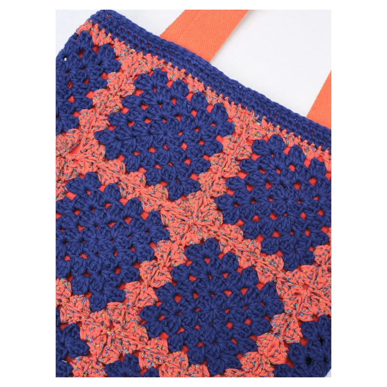 May include: A crocheted tote bag with a granny square pattern in navy blue and coral. The bag has orange straps and a matching navy blue border. The crochet work is detailed, showcasing the texture and design of the bag.