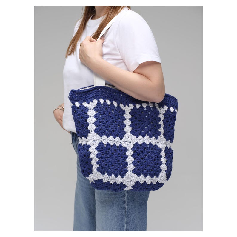 May include: A navy blue and white crochet tote bag with a geometric pattern. The bag has a rectangular shape with a white strap and a zipper closure. The crochet design features a grid-like pattern.