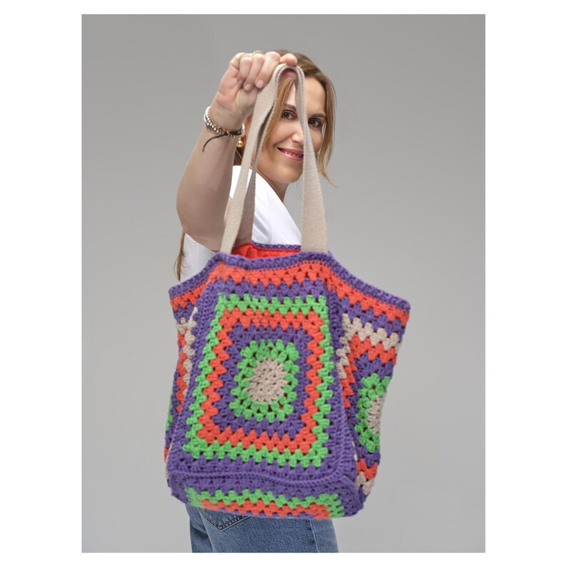Puede incluir: Un bolso tote tejido a ganchillo, colorido, con un patr&oacute;n geom&eacute;trico. El bolso presenta un dise&ntilde;o cuadrado con colores alternos de morado, verde, naranja y beige. Las asas son de color beige neutro. El bolso es sostenido por una persona.