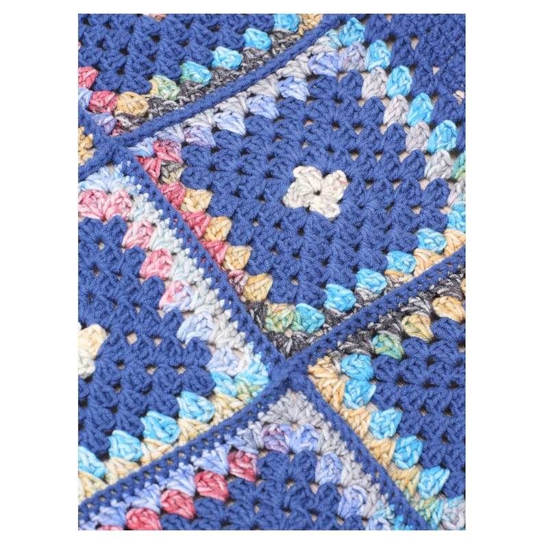 Puede incluir: Primer plano de una manta azul tejida a ganchillo con un dise&ntilde;o de cuadrados de la abuela. La manta presenta cuadrados en tonos azules, con acentos coloridos de rosa, amarillo y gris. La textura del hilo es visible, creando una sensaci&oacute;n acogedora y hecha a mano.