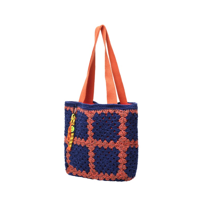 May include: A handmade crochet tote bag in navy blue and coral. The bag features a square pattern design and orange handles. A small, colorful charm hangs from the side. This stylish bag is perfect for carrying everyday essentials.