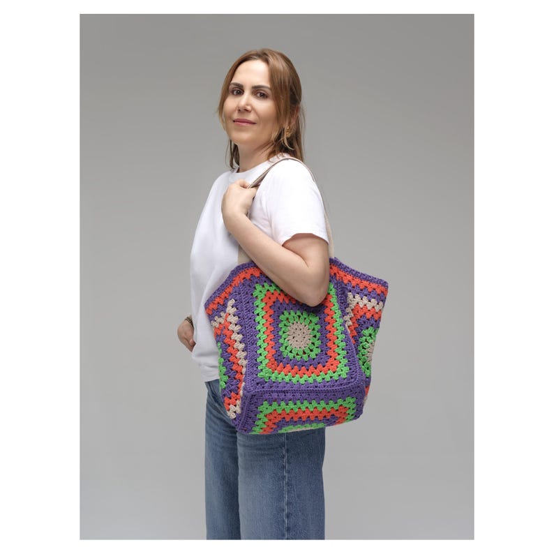 Puede incluir: Un bolso tote de ganchillo colorido con un patr&oacute;n geom&eacute;trico en tonos morado, verde, naranja y beige. El bolso tiene forma rectangular y asas marrones. El bolso lo lleva una persona que viste una camiseta blanca y vaqueros azules.