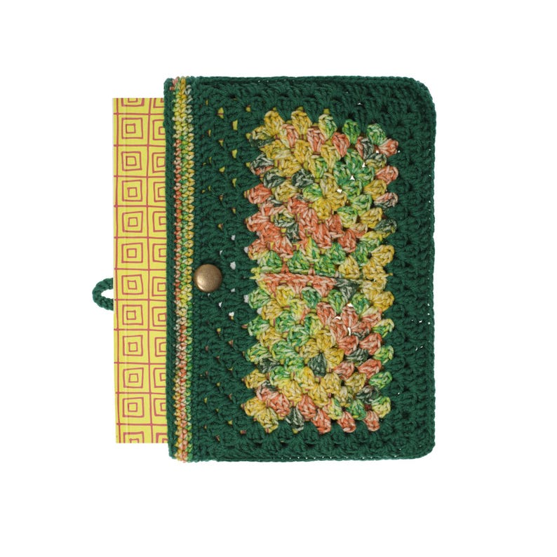 Puede incluir: Una funda rectangular para cuaderno con exterior de ganchillo verde oscuro y dos paneles centrales de hilo jaspeado en amarillo, naranja y verde. Un &uacute;nico bot&oacute;n de presi&oacute;n met&aacute;lico asegura la funda, que envuelve un cuaderno amarillo con cuadr&iacute;cula roja.