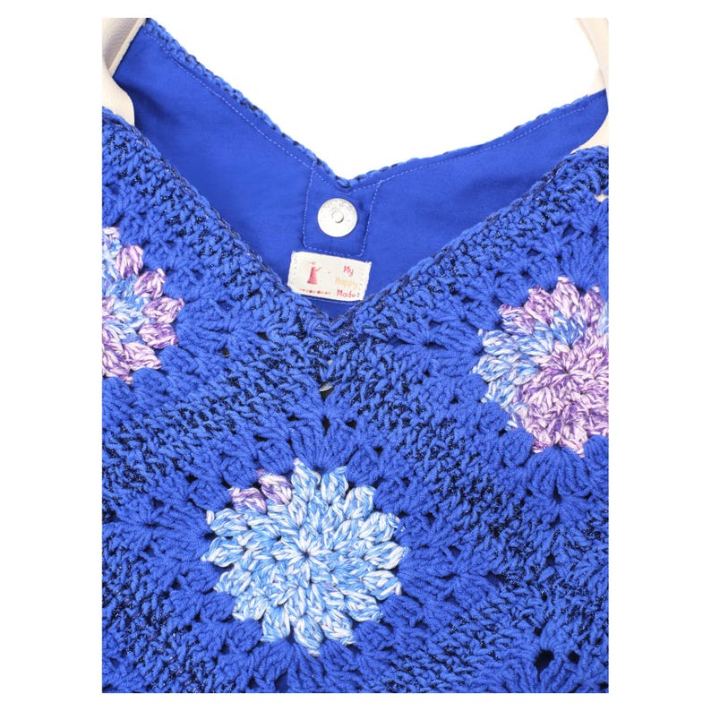 May include: A blue crochet bag with a floral pattern in shades of blue, purple, and white. The bag has a solid blue lining and a white strap. A small tag is visible near the top.