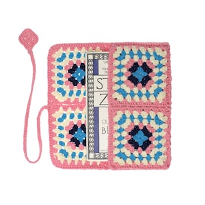 K&ouml;nnte beinhalten: Ein rechteckiger H&auml;kel-Buchumschlag mit Granny-Square-Muster in Rosa, Creme, Hellblau und Dunkelblau. Der Umschlag enth&auml;lt ein befestigtes rosa H&auml;kel-Lesezeichen mit rautenf&ouml;rmigem Ende. Ein Buch mit sichtbarem Text befindet sich darin.