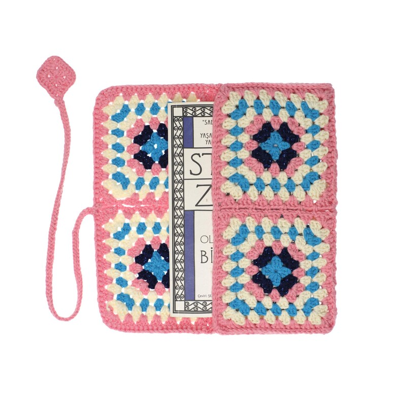 K&ouml;nnte beinhalten: Ein rechteckiger H&auml;kel-Buchumschlag mit Granny-Square-Muster in Rosa, Creme, Hellblau und Dunkelblau. Der Umschlag enth&auml;lt ein befestigtes rosa H&auml;kel-Lesezeichen mit rautenf&ouml;rmigem Ende. Ein Buch mit sichtbarem Text befindet sich darin.