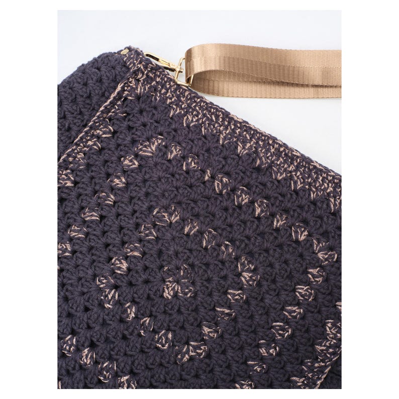 May include: A dark charcoal gray crochet bag featuring a repeating square pattern with subtle metallic gold accents. The bag includes a wide, smooth beige nylon strap attached with a gold-toned metal clasp, showcasing a textured yarn construction.