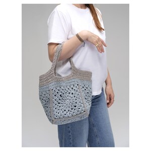 May include: A handmade crochet tote bag in shades of gray and light blue. The bag features a geometric pattern and sturdy handles. The bag is held by a person wearing a white t-shirt and blue jeans.