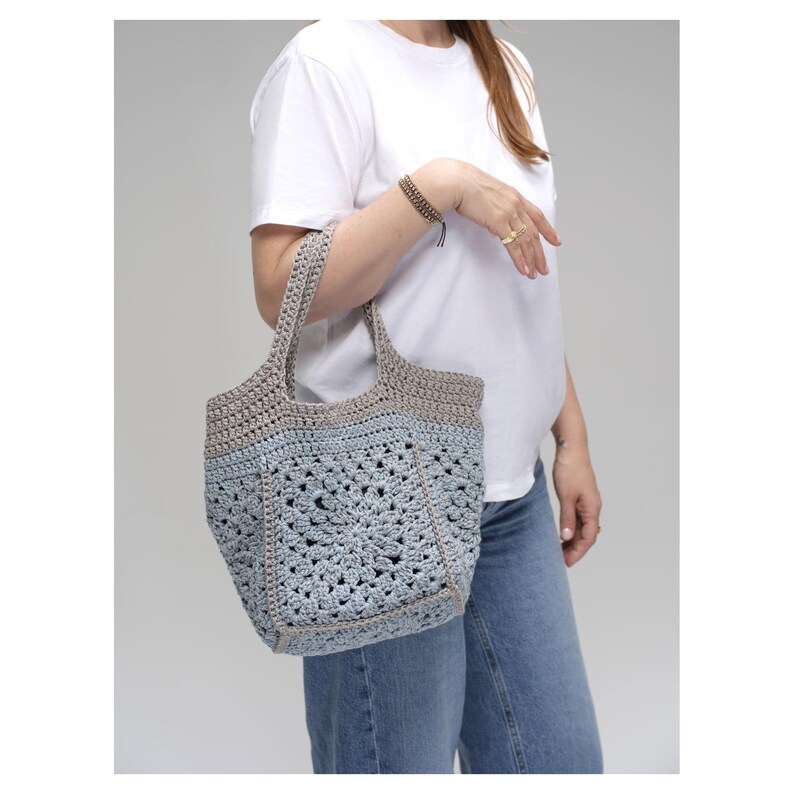 May include: A handmade crochet tote bag in shades of gray and light blue. The bag features a geometric pattern and sturdy handles. The bag is held by a person wearing a white t-shirt and blue jeans.