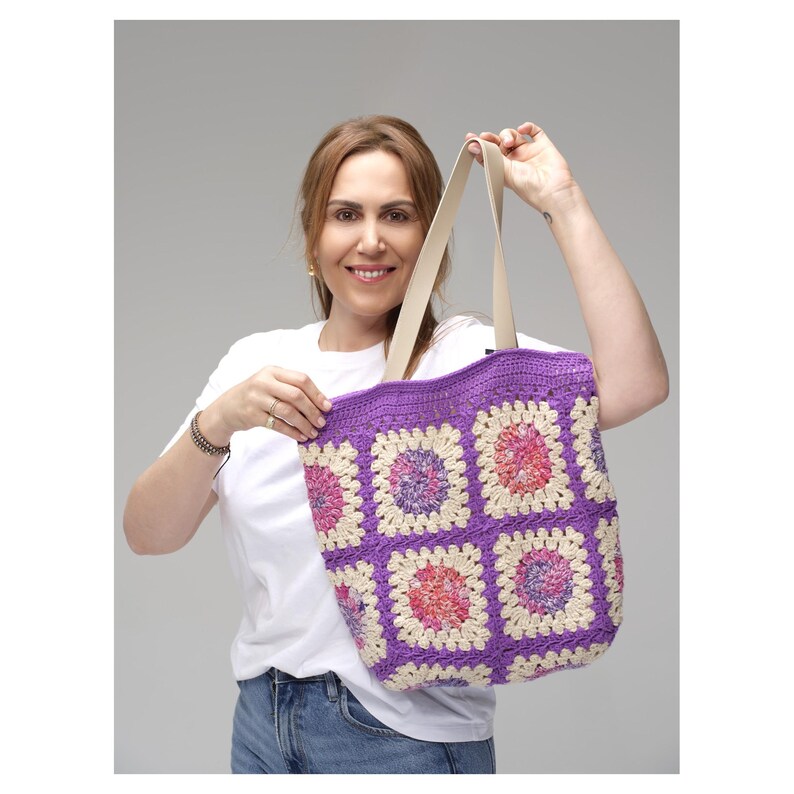 May include: A handmade crochet tote bag with a vibrant purple border and cream-colored straps. The bag features a granny square design with squares of purple, pink, and cream. This stylish tote is perfect for carrying everyday essentials.