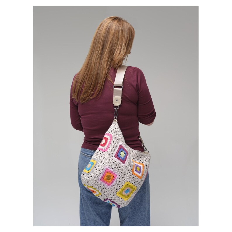 May include: A cream crochet shoulder bag with colourful square patterns. The bag has a long, adjustable strap and is worn over the shoulder. The squares feature various colours, including pink, yellow, blue, and green.