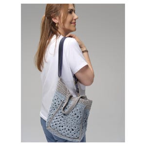 May include: A handmade crochet tote bag in shades of blue and gray, with a dark blue leather strap. The bag has a textured, open weave design and a rectangular shape. The strap is adjustable and attached with gold-colored hardware.
