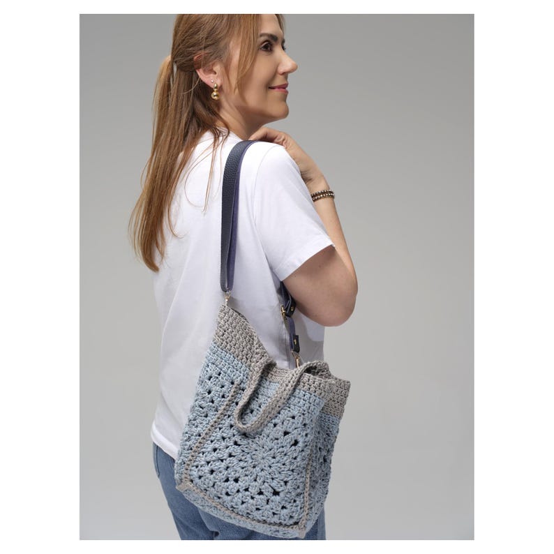 May include: A handmade crochet tote bag in shades of blue and gray, with a dark blue leather strap. The bag has a textured, open weave design and a rectangular shape. The strap is adjustable and attached with gold-colored hardware.