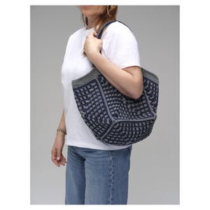 May include: A navy and grey crocheted tote bag with a textured pattern. The bag has a rectangular shape with a grey border and handles. The bag is held by a person wearing a white t-shirt and blue jeans.