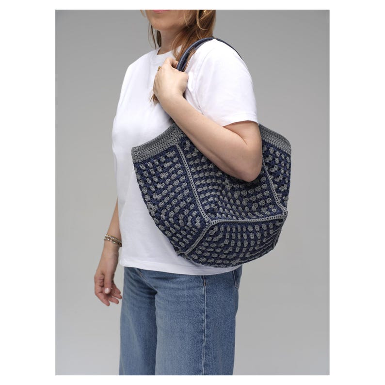May include: A navy and grey crocheted tote bag with a textured pattern. The bag has a rectangular shape with a grey border and handles. The bag is held by a person wearing a white t-shirt and blue jeans.