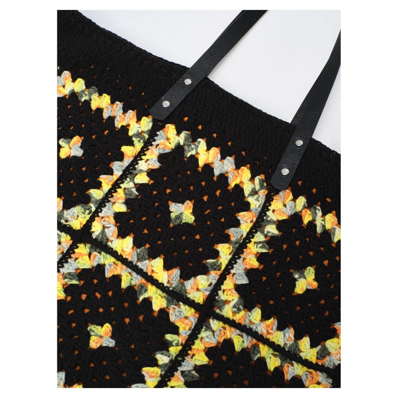 Puede incluir: Bolso tote de crochet negro con un colorido estampado floral en amarillo, naranja y gris. El bolso tiene correas de cuero negro sujetas con remaches plateados. Un accesorio elegante para llevar objetos.