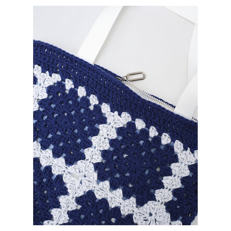 May include: A close-up of a navy blue and white crocheted tote bag. The bag features a geometric pattern and a zipper closure. White straps are visible, and the bag is set against a white background. The bag is a stylish accessory.