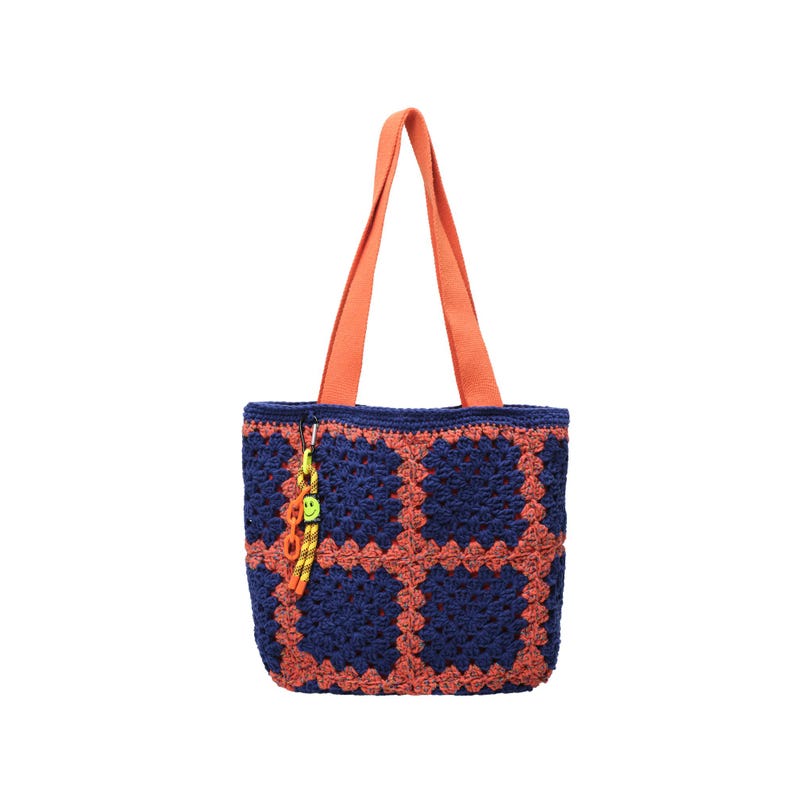 May include: A handmade tote bag with a crochet design. The bag features a navy blue and orange color scheme, with orange handles and a matching crochet pattern. A small charm with a smiley face hangs from the bag.