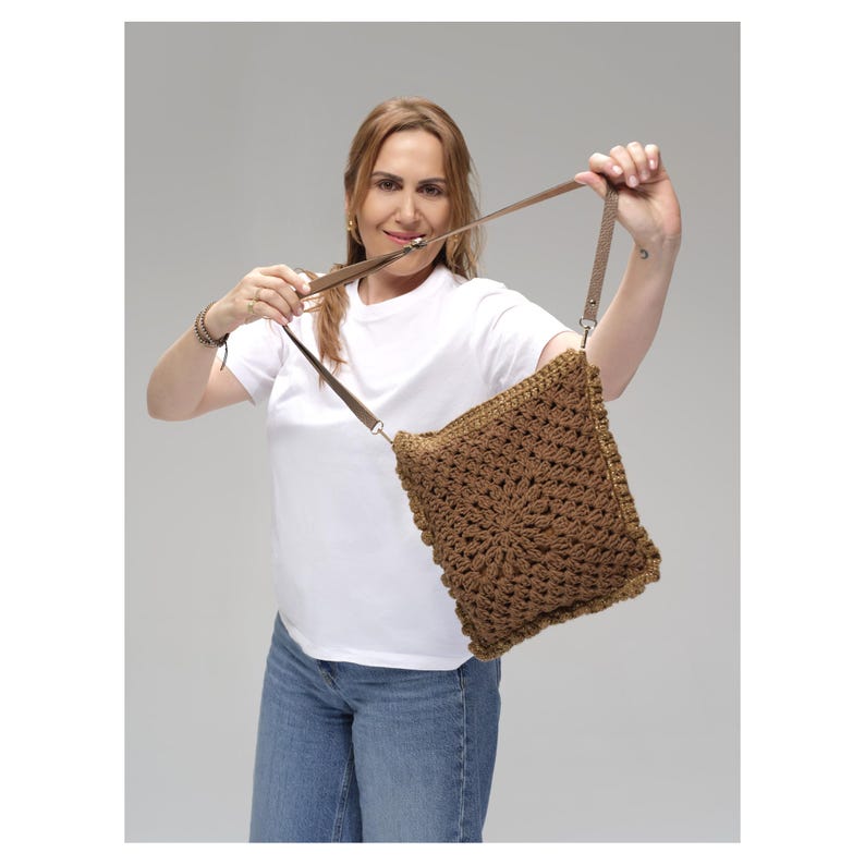 May include: A square-shaped brown crochet shoulder bag featuring a detailed circular floral pattern in the center. The bag is constructed with thick yarn and includes a matching brown adjustable strap attached with silver-toned metal hardware.
