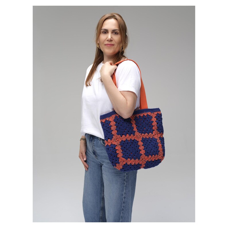 May include: A blue and orange crochet tote bag with a square pattern. The bag has a solid blue base with orange squares and an orange strap. The bag is carried over the shoulder.