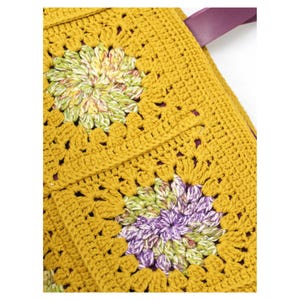 Puede incluir: Bolso tote de crochet amarillo mostaza con dise&ntilde;o floral de granny square. El bolso presenta una correa de cuero morado. Los cuadrados florales tienen detalles verdes, morados y blancos. El bolso es hecho a mano.