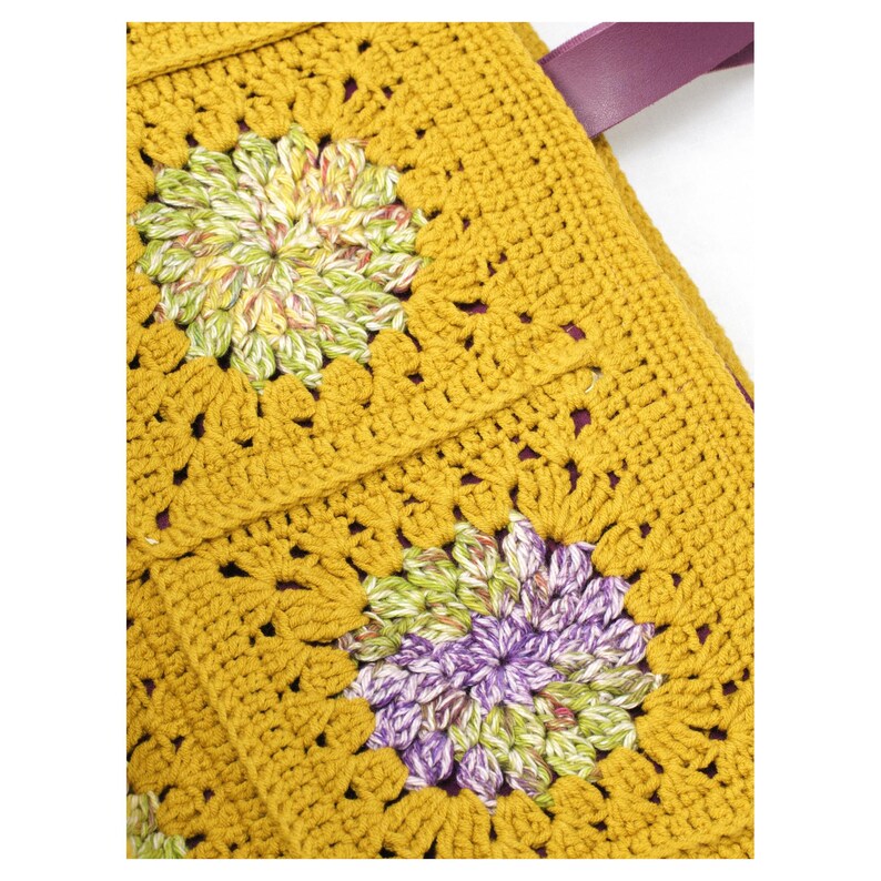 Puede incluir: Bolso tote de crochet amarillo mostaza con dise&ntilde;o floral de granny square. El bolso presenta una correa de cuero morado. Los cuadrados florales tienen detalles verdes, morados y blancos. El bolso es hecho a mano.