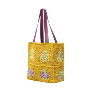 Puede incluir: Un bolso tote de crochet amarillo con asas de cuero morado. El bolso presenta un dise&ntilde;o de cuadrados de la abuela con motivos florales verdes y morados. El bolso tiene forma cuadrada y es adecuado para transportar varios art&iacute;culos.
