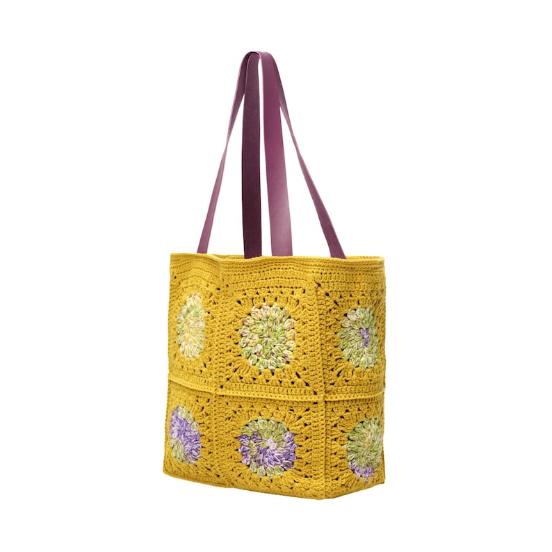 Puede incluir: Un bolso tote de crochet amarillo con asas de cuero morado. El bolso presenta un dise&ntilde;o de cuadrados de la abuela con motivos florales verdes y morados. El bolso tiene forma cuadrada y es adecuado para transportar varios art&iacute;culos.