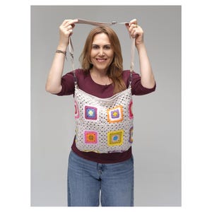 May include: A cream-coloured crochet bag with colourful square patterns. The bag has a long strap. The squares feature bright colours like pink, yellow, blue, and orange. The bag is held up by a person.