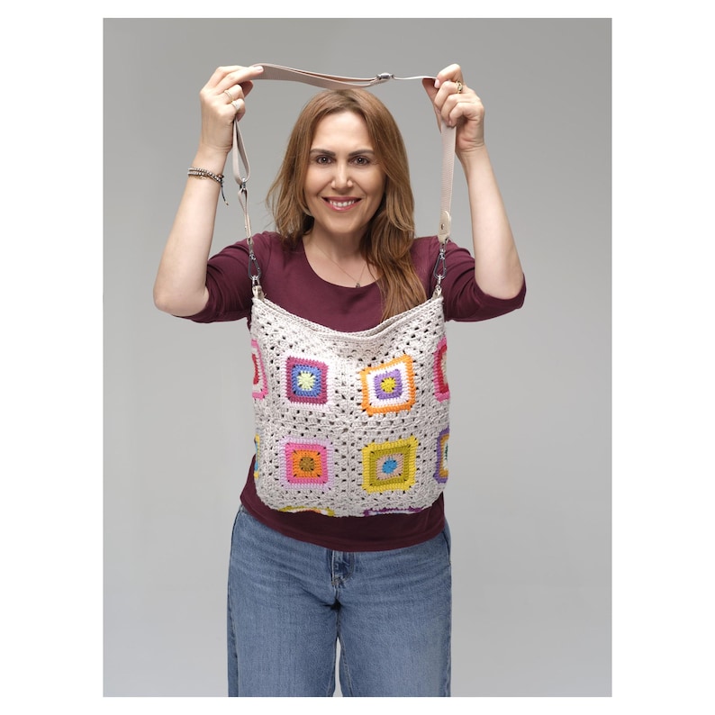 May include: A cream-coloured crochet bag with colourful square patterns. The bag has a long strap. The squares feature bright colours like pink, yellow, blue, and orange. The bag is held up by a person.