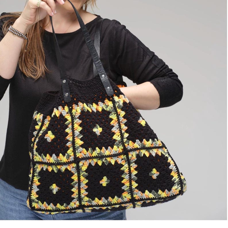 Puede incluir: Bolso tote de crochet negro con un patr&oacute;n geom&eacute;trico en amarillo, gris y naranja. El bolso tiene asas de cuero negro. El bolso es un art&iacute;culo hecho a mano. El bolso lo sostiene una persona.