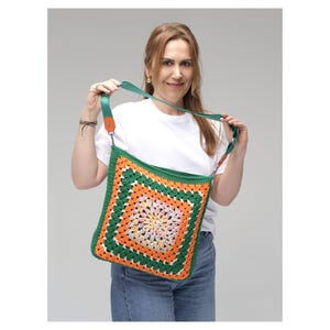 K&ouml;nnte beinhalten: Handgefertigte geh&auml;kelte Umh&auml;ngetasche mit quadratischem Design. Die Tasche hat ein gr&uuml;nes, orangefarbenes und cremefarbenes Farbschema. Der Riemen ist gr&uuml;n mit orangefarbenen Akzenten. Die Tasche wird von einer Person gehalten, die ein wei&szlig;es Hemd und blaue Jeans tr&auml;gt.