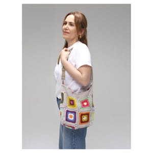 K&ouml;nnte beinhalten: Eine beige H&auml;keltasche mit bunten quadratischen Mustern. Die Tasche hat einen langen Riemen und wird &uuml;ber der Schulter getragen. Die Quadrate sind in verschiedenen Farben, darunter Rosa, Gelb, Gr&uuml;n und Lila. Die Tasche ist handgefertigt.