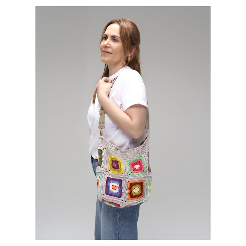 K&ouml;nnte beinhalten: Eine beige H&auml;keltasche mit bunten quadratischen Mustern. Die Tasche hat einen langen Riemen und wird &uuml;ber der Schulter getragen. Die Quadrate sind in verschiedenen Farben, darunter Rosa, Gelb, Gr&uuml;n und Lila. Die Tasche ist handgefertigt.