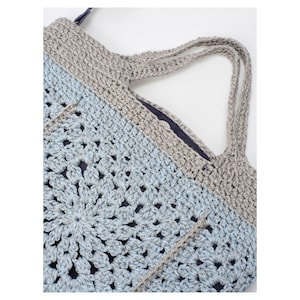 May include: A handmade crochet tote bag in shades of light blue and gray. The bag features a floral pattern on the front and a solid gray top with matching handles. The interior is lined with a dark blue fabric.