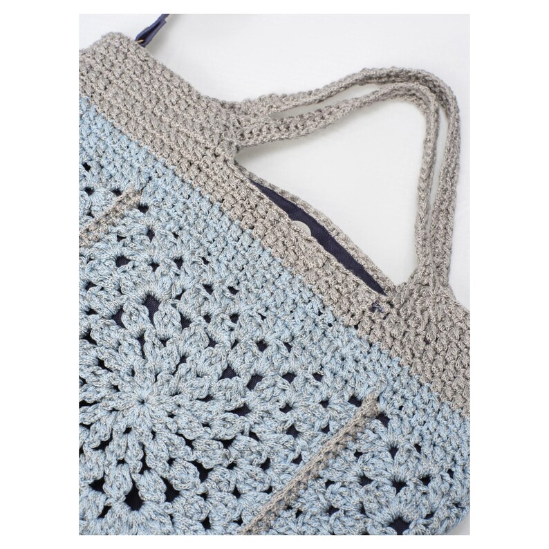 May include: A handmade crochet tote bag in shades of light blue and gray. The bag features a floral pattern on the front and a solid gray top with matching handles. The interior is lined with a dark blue fabric.