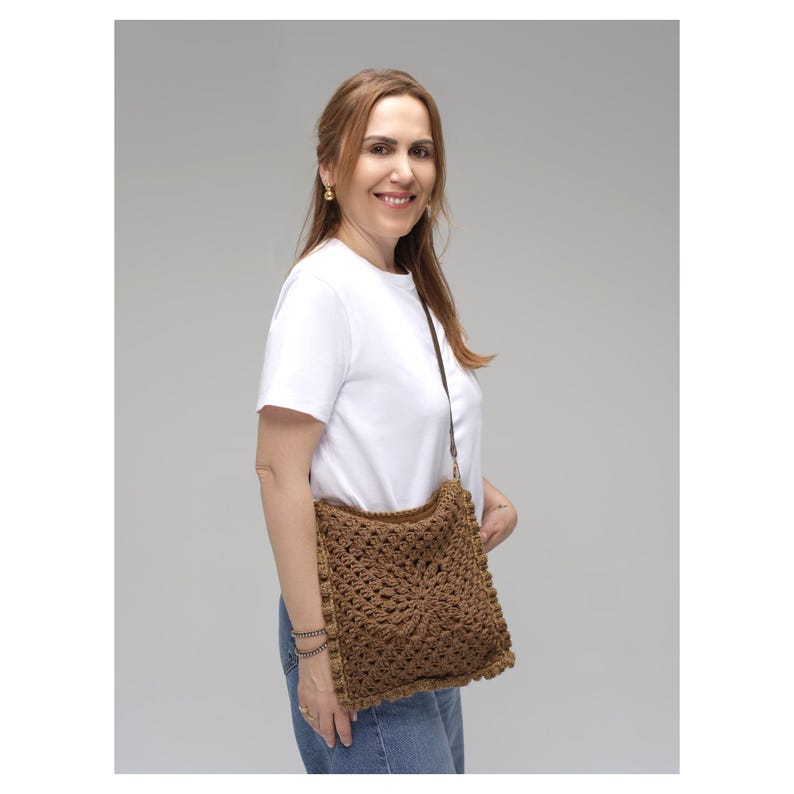 May include: A brown crochet crossbody bag featuring a square-shaped design with an intricate floral-inspired pattern and a textured border. The bag is attached to a thin, dark brown strap that rests over the shoulder, providing a functional accessory.