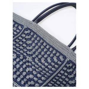 May include: A crocheted tote bag in navy blue and grey. The bag has a geometric pattern, a grey border, and navy blue handles. The bag is made from a soft, textured material, perfect for carrying items.