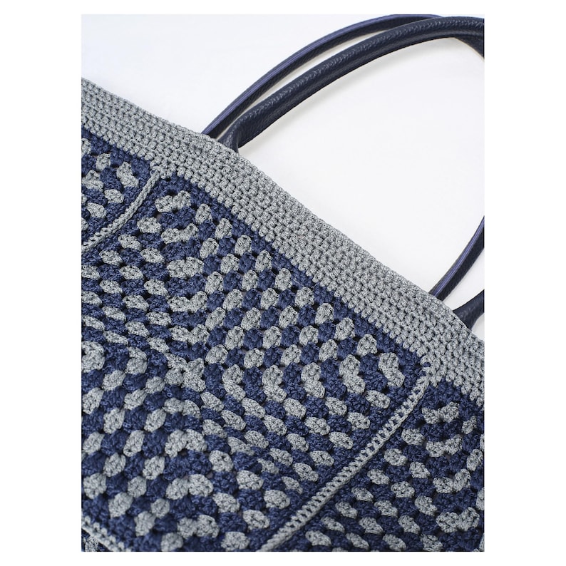 May include: A crocheted tote bag in navy blue and grey. The bag has a geometric pattern, a grey border, and navy blue handles. The bag is made from a soft, textured material, perfect for carrying items.
