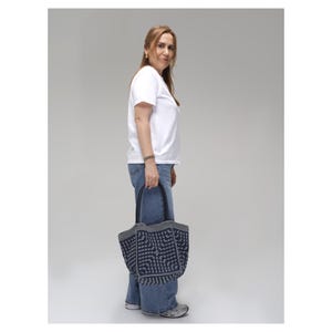 May include: A woman in a white t-shirt and blue jeans carries a navy tote bag with a geometric design. The bag has grey trim and black handles. The woman is standing against a plain grey backdrop.
