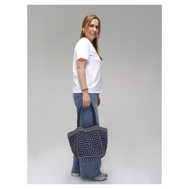 May include: A woman in a white t-shirt and blue jeans carries a navy tote bag with a geometric design. The bag has grey trim and black handles. The woman is standing against a plain grey backdrop.