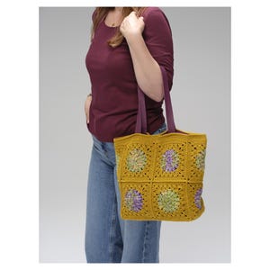 Puede incluir: Bolso tote de crochet amarillo mostaza con detalles florales morados y verdes. El bolso presenta un dise&ntilde;o de patr&oacute;n cuadrado y correas moradas. El bolso es llevado por una persona que viste una camisa burdeos y vaqueros azules.